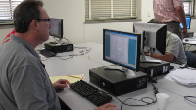 A man sits at a computer looking at a screen with irrigation plans on it. There are other computers in the room and students taking the computer aided software design course.