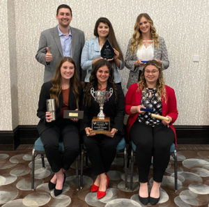 Two rows of six people, one set of three stand, the other set of three sit with trophies and other winnings as the champion 2022 Poultry Judging Team