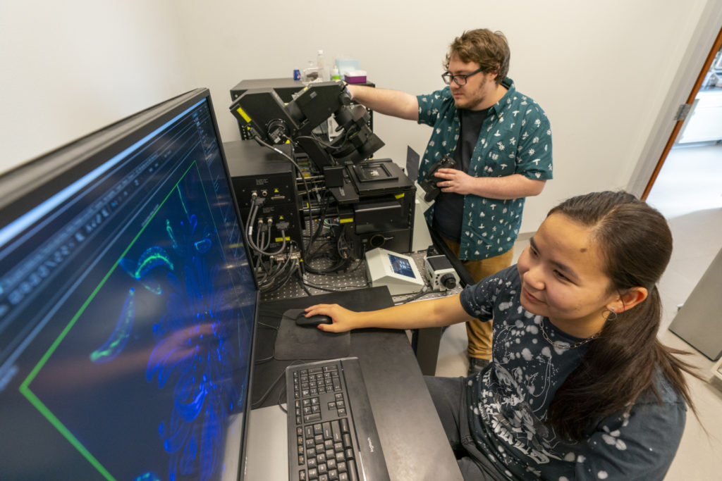 A woman, Mary Cowser, sits at a computer in the foreground and a man, Oli Bedsole, stands using the confocal microscope in the background.