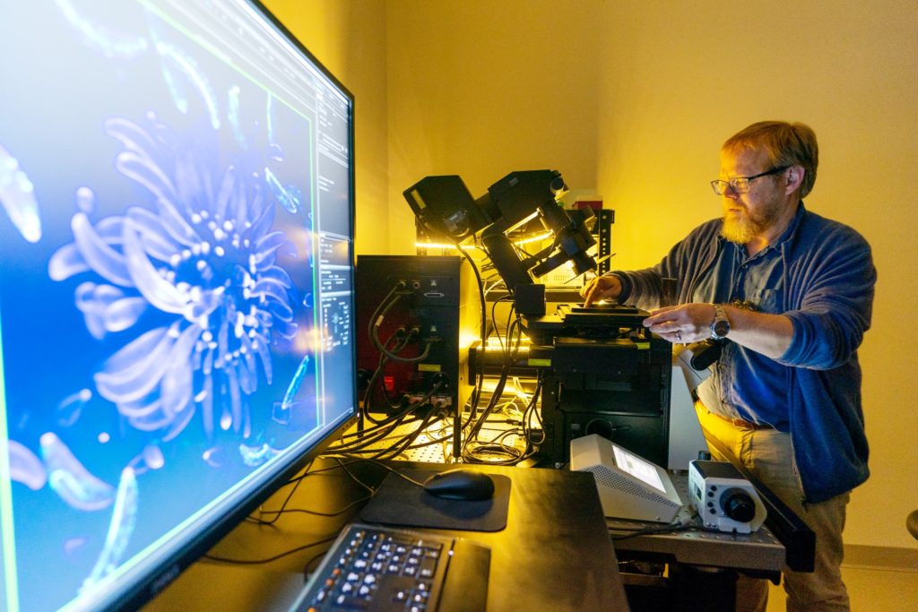 A man, Brian Shaw, Ph.D., stands behind the large confocal microscope, with an Image from microscope enlarged on nearby monitor and a yellow light bouncing back off on Shaw's face.