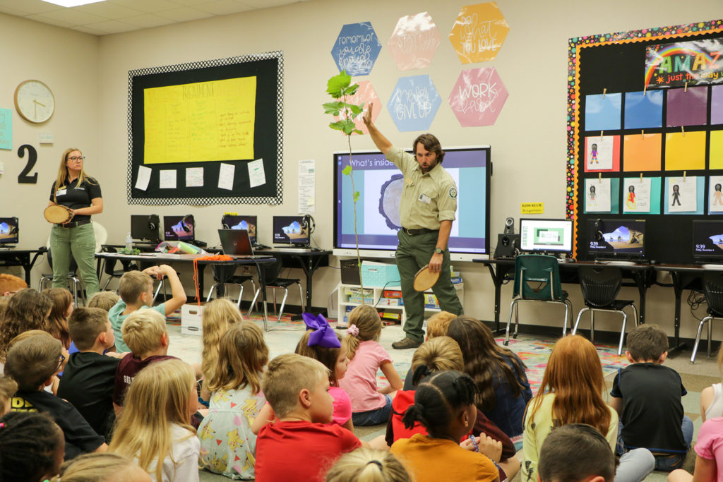 A man stands in the front of a classroom reaching up to the top of a potted tree with a bunch of small children sitting on the floor with their backs to us.