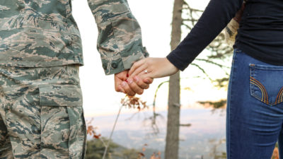 A man in a camouflaged military uniform holds the hand of a woman in jeans and a black long sleeve shirt. We see them from behind from the waist down. They are standing in a wooded area.