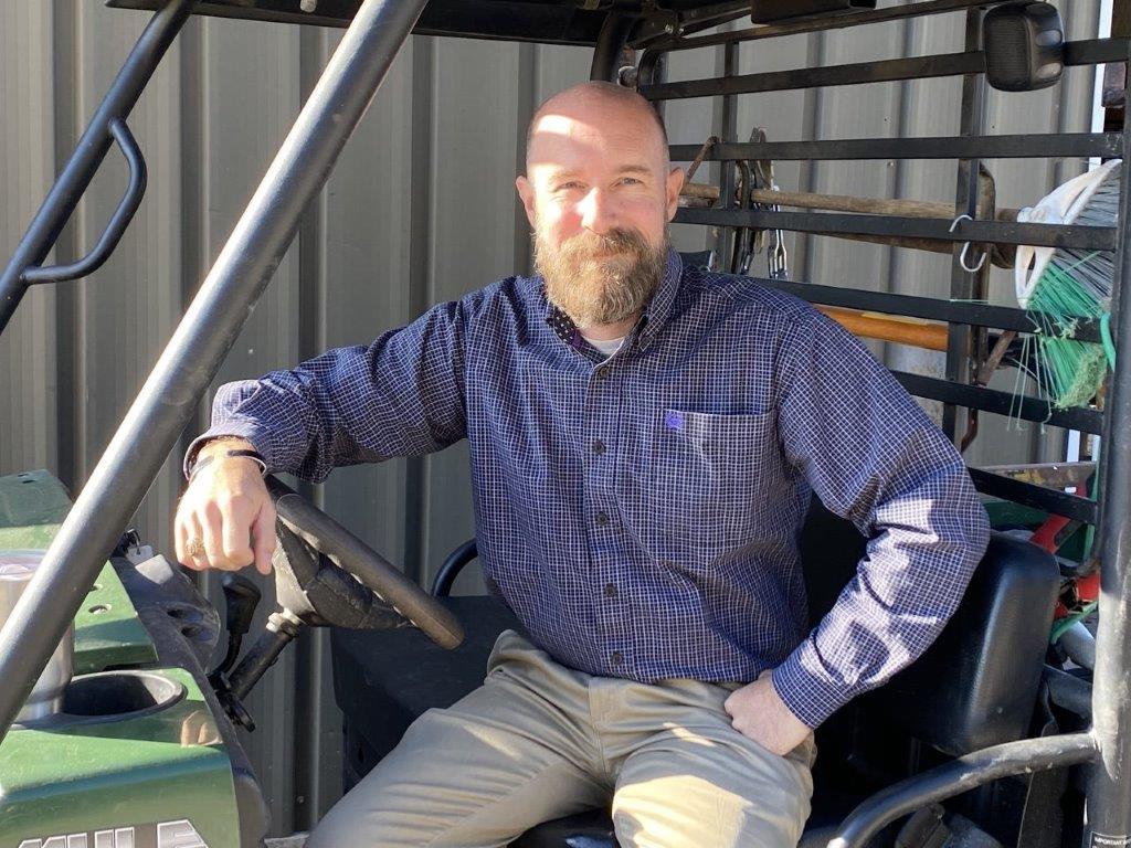 A bearded man, Greg Wilson, sits in farm vehicle and smiles at the camera.