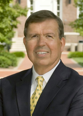The head and shoulders of a white man, Donald Sparks, in a dark suite with a yellow tie with trees and a building in the background.