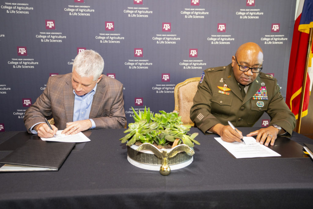 Two men, one in a military uniform, sit at a table signing a memorandum in front of a College of Agriculture and Life Sciences backdrop.