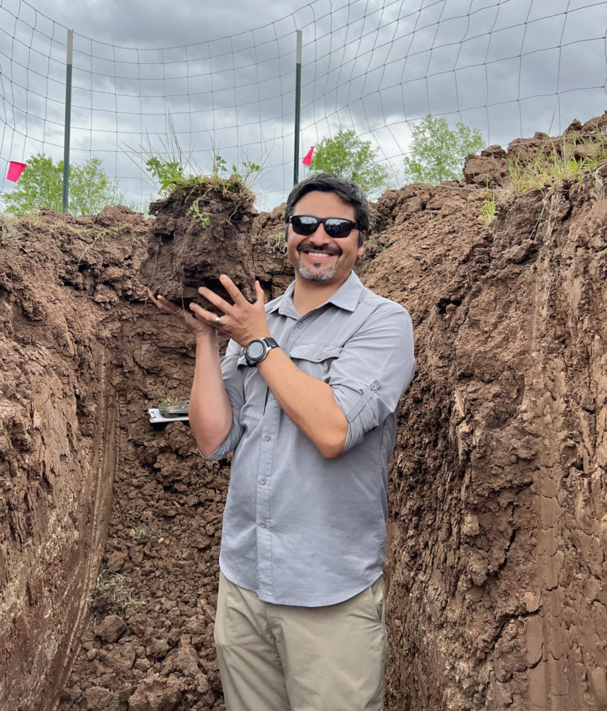 Felipe Aburto stands in a ditch with dirt walls as high as his head and he holds a chunk of soil with green plants growing out of it 