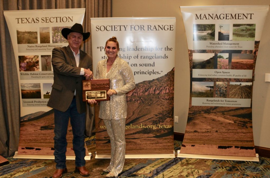 A man wearing a cowboy hat presents an award plaque to a woman standing to his left. The two individuals stand in front of three tall banners. 