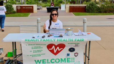 Female volunteer showing "Gig 'em" sign sits at REACH health fair table in the Leach Teaching Gardens.