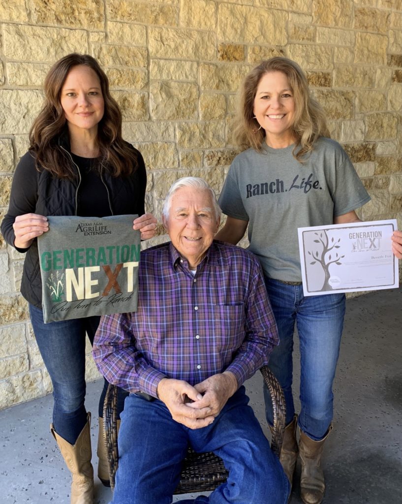 Two women stand to showcase their Generation Next T-shirt and certificate of completion. An older gentleman is seated between them. 
