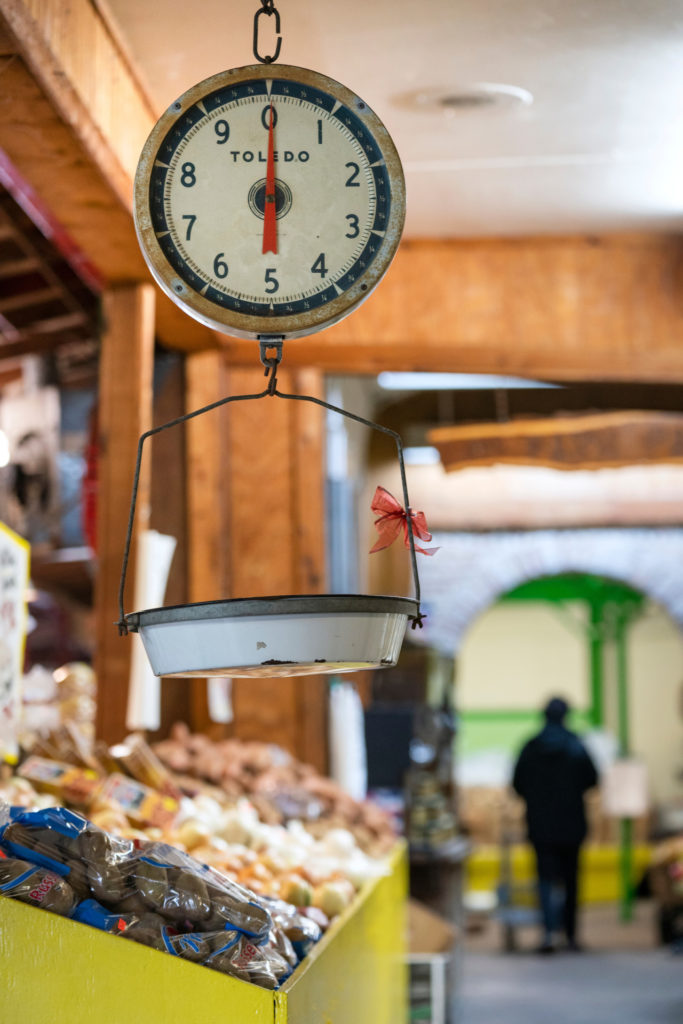 A hanging produce scale above bins of vegetables in a framers market type of setting.