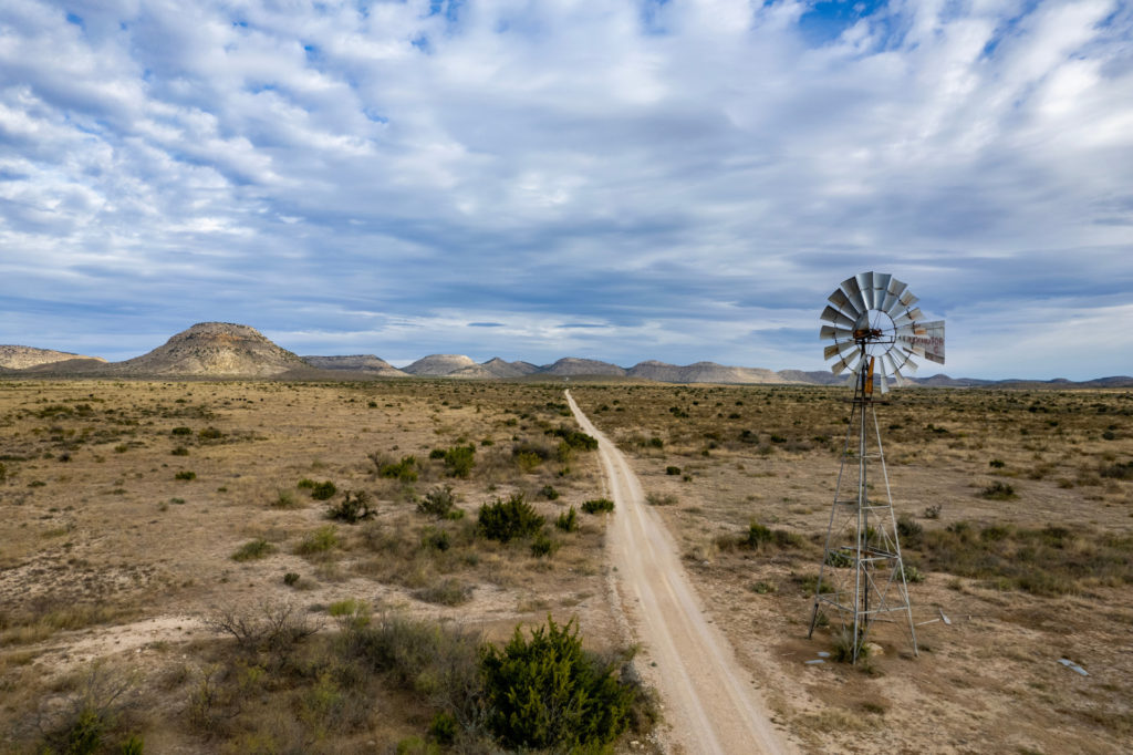 A wide open West Texas scene with a dirt road running through the middle of ranchland under a blue sky with clouds.