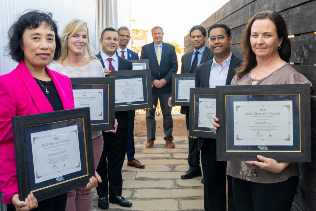 Eight people holding awards certificates standing in a V shape on a flagstone patio smiling 