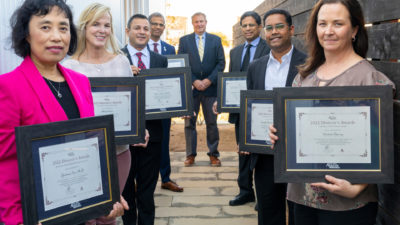 Eight people holding awards certificates standing in a V shape on a flagstone patio smiling