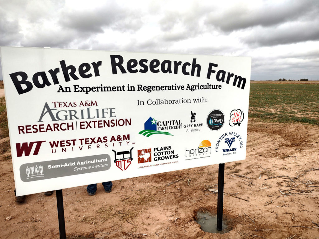 A large sign in a sparsely cropped field reads Barker Research Farm, an experiment in regenerative agriculture with many collaborator logos on it.