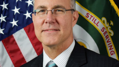 A portrait of Mark Overberg, retired US Army, wearing a dark suit with the US and a military flag behind him.