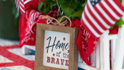 A sign that reads Home of the Brve in a centerpiece with red flowers, green leaves and two American flags.