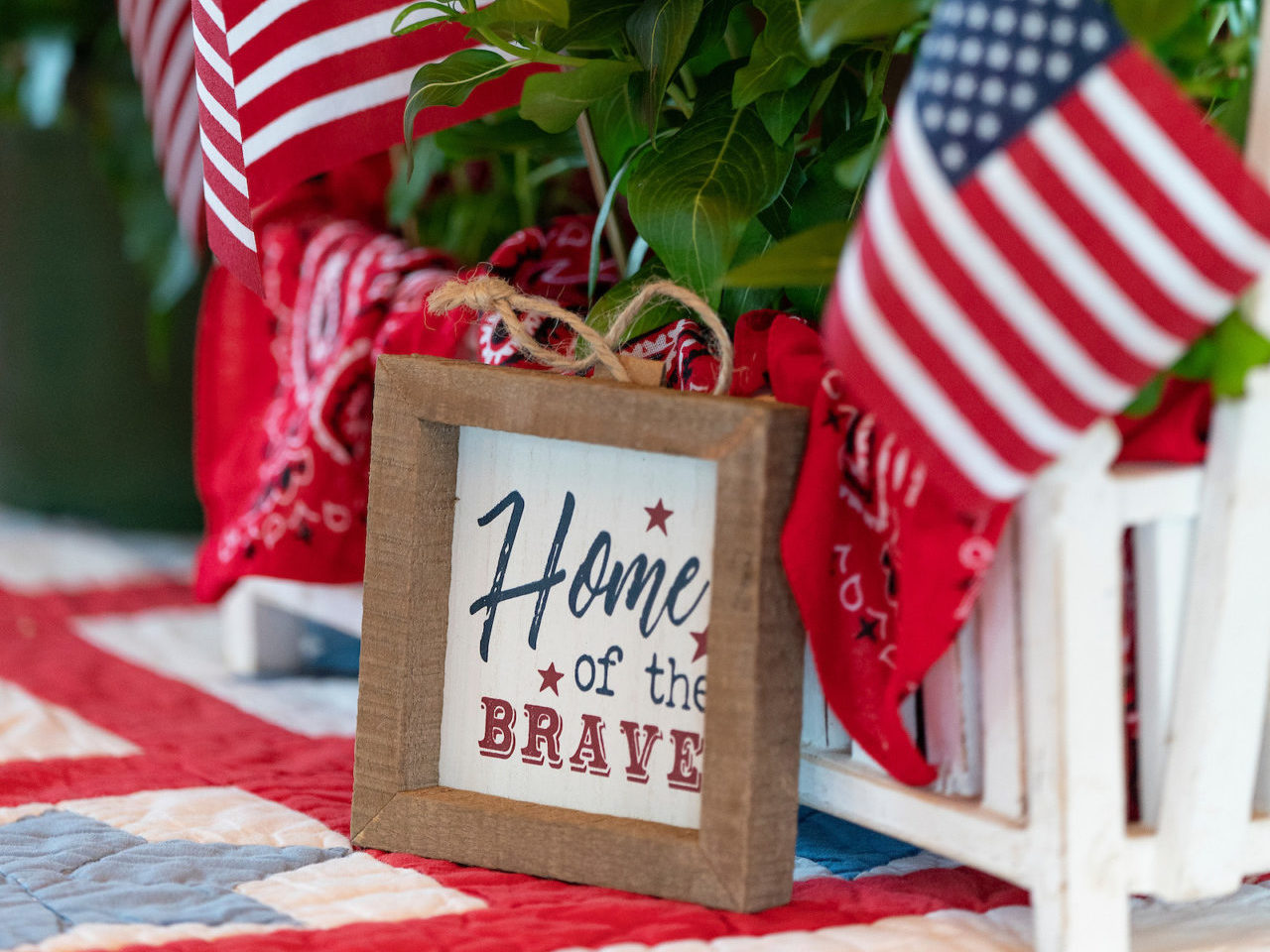 A sign that reads Home of the Brave in a plant centerpiece on a quilt covered table with red flowers, green leaves and two American flags.