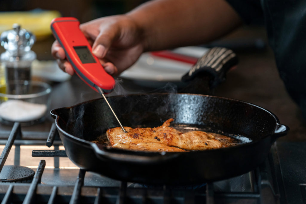 A piece of chicken in a skillet with a temperature thermometer being inserted in it for food safety.
