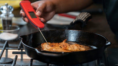 A piece of chicken in a skillet with a temperature thermometer being inserted in it for food safety.