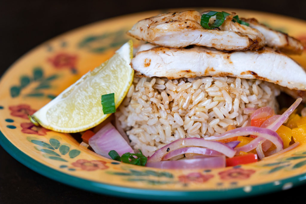 A grille chicken breast atop rice, onion and peppers on a colorful yellow floral plate. 