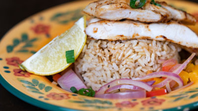 A grille chicken breast atop rice, onion and peppers on a colorful yellow floral plate.