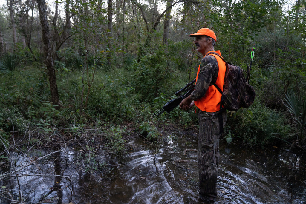 A bow hunter stands in a small pond in a wooded area. He wears a bright orange vest and hat over camo and holds his bow.