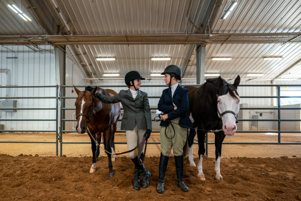 Two female 4-H riders in dressage gear standing next to one another along with their horses 