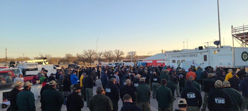 Large group of firefighters assemble in front of Texas A&M Forest Service trailer for a morning briefing.