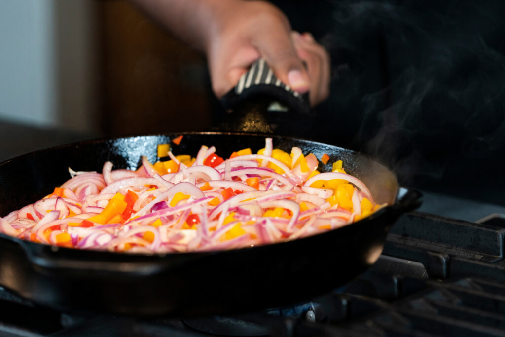 A healthy cooking demonstration. A hand holds a skillet on a stove that is full of red onions and yellow and red peppers.