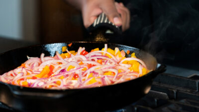 A cooking demonstration. A hand holds a skillet on a stove that is full of red onions and yellow and red peppers.