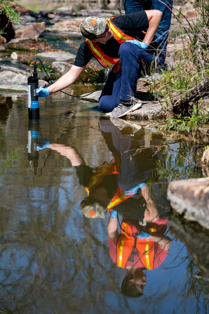 Two students and their reflection in the water as they take a water sample in White Creek at the Leach Teaching Gardens at Texas A&M University.