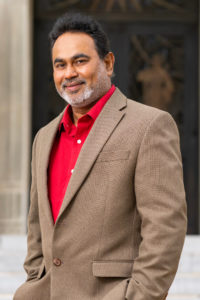 A man in a brown suit with a bright red shirt Dr. Sandun Fernando 