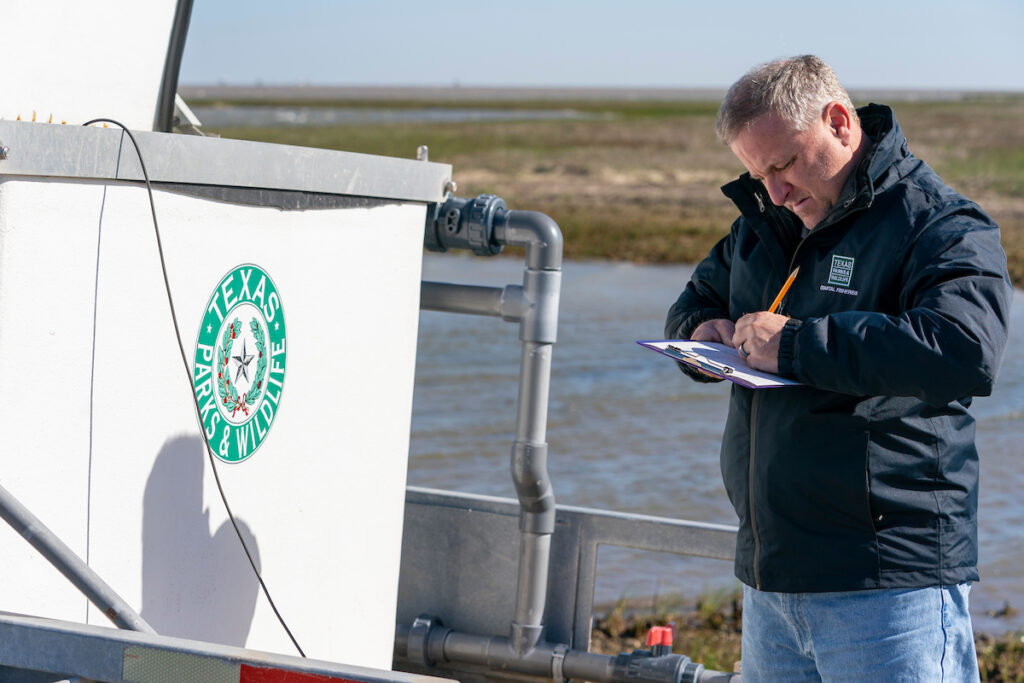 A man in a Texas Parks and Wildlife jacket takes notes while standing next to a body of water. 