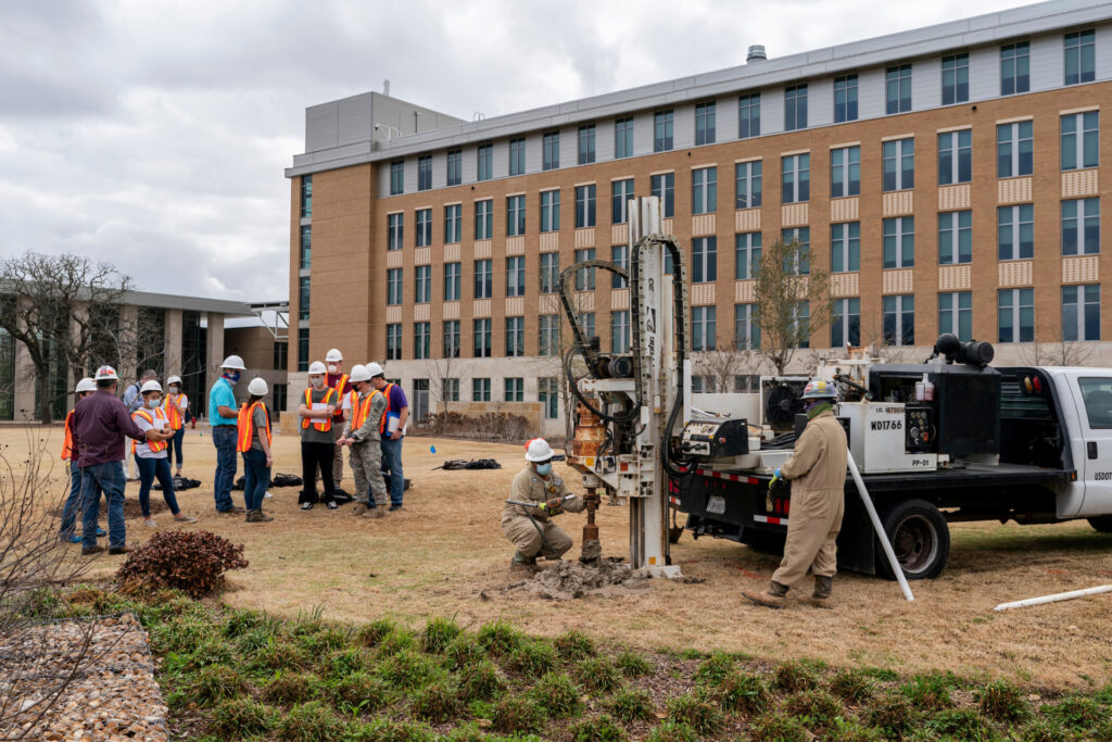 Group of bioenvironmental sciences students in hard hats watch two workers behind a large truck and mechanical rig install a water well.