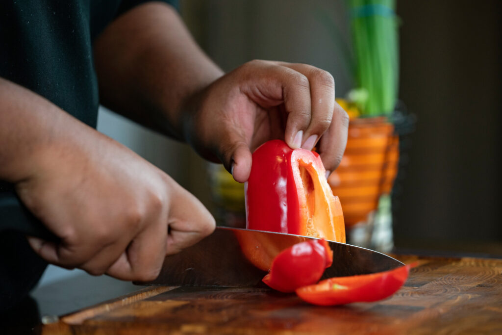 A pair of hands holds and chops a red bell pepper on a dark wood cutting board