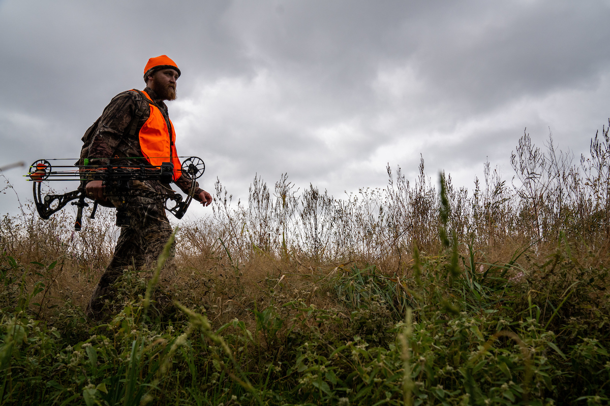 A man with a compound bow walks through a field.