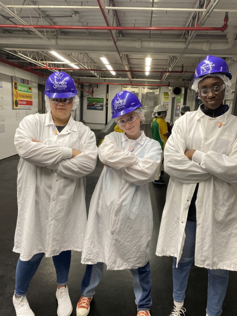 Three young women pose in hard hats, hair nets and coats before touring a frozen foods plant.