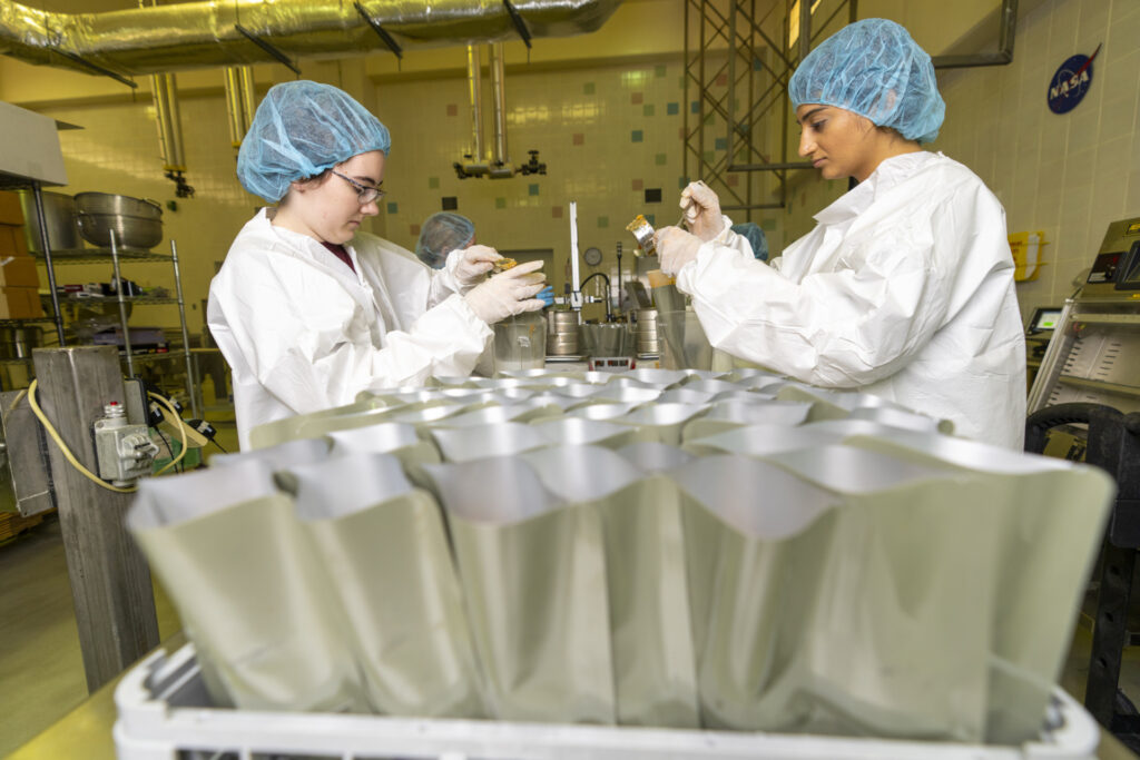 Space food facility workers with open MRE-type pouches in foreground.