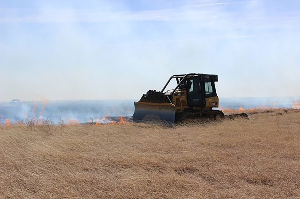 a motor-grader pushes dirt over small  flames in a field of dried grass