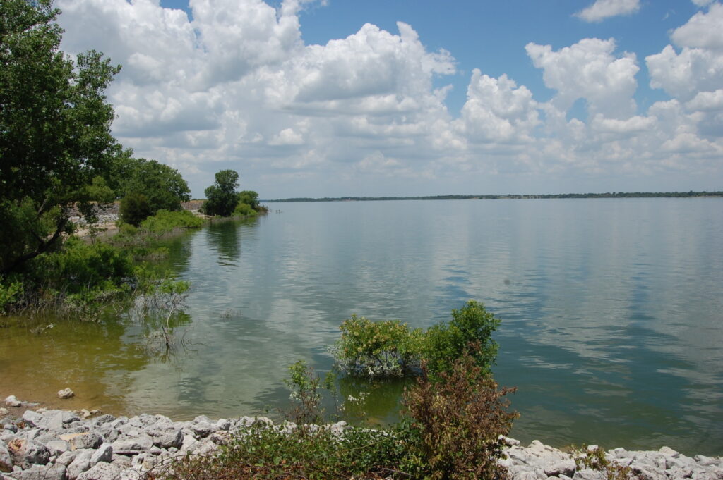 a big body of water with one tree-lined shore showing under blue cloud-filled skies.