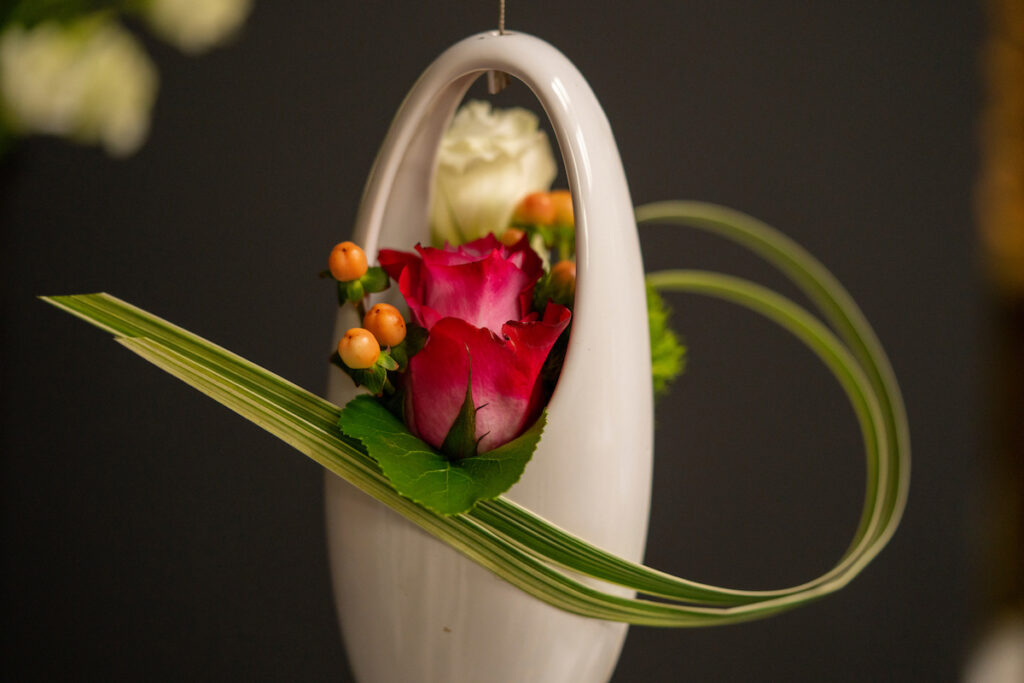 A floral design that appears to be inspired by the art of Ikebana. A hanging white porcelain vase holds two roses while a long leaf curves around it. The career day at Texas A&M will cover this and other horticulture topics and career options within the horticulture sciences.