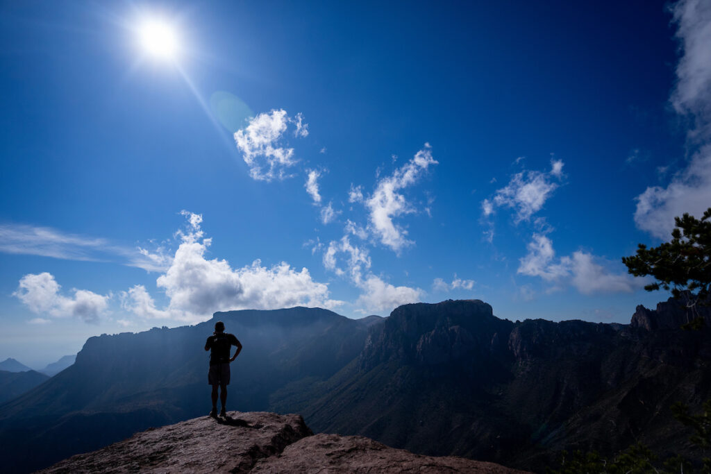 A man stands atop a mountain in Big Bend in Texas gazing toward the horizon. The sky is bright blue with a few white clouds.