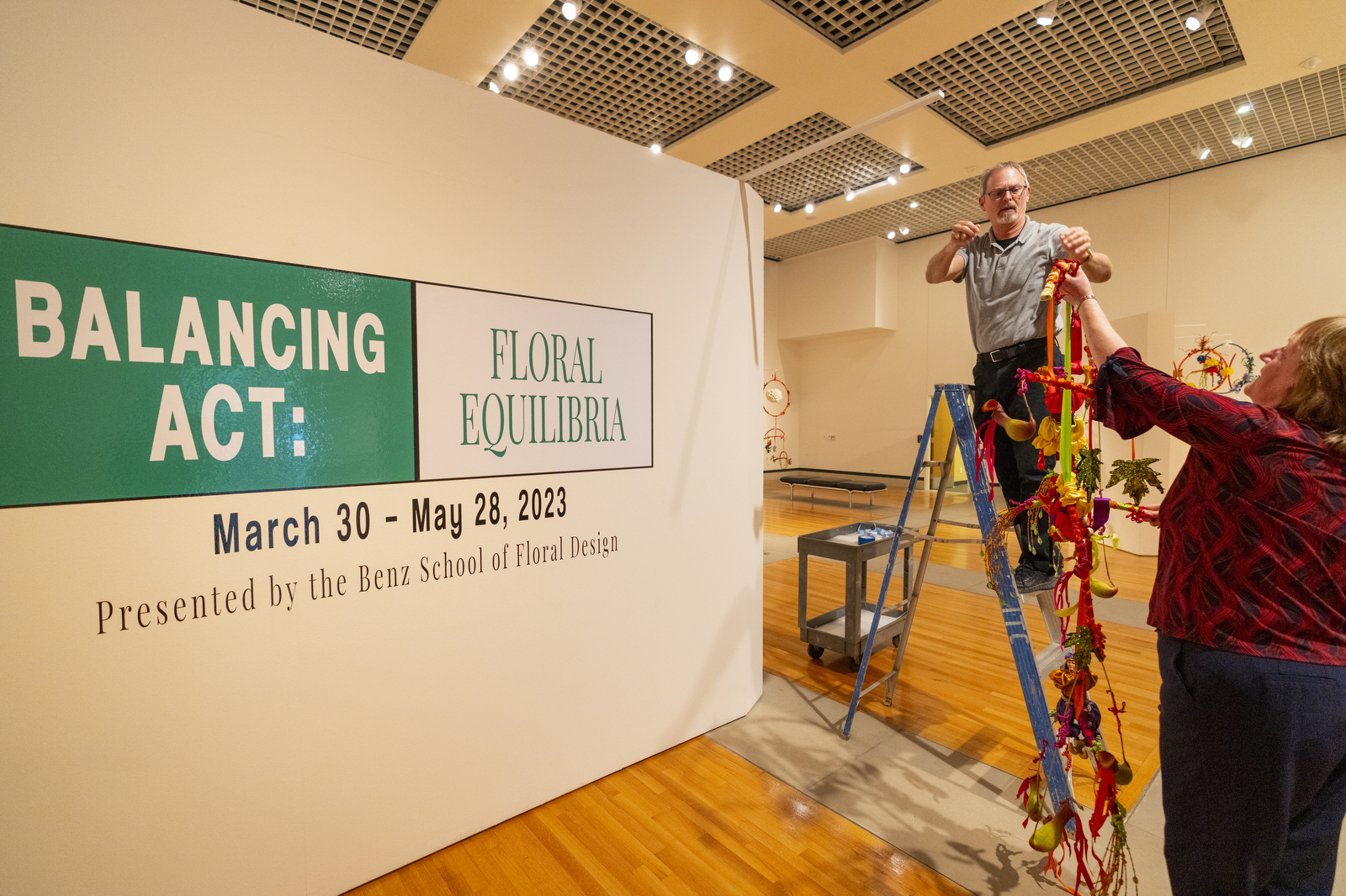 William McKinley hangs a floral sculpture in front of a sign reading Balancing Act: FLoral Equlibria. March 30-May28, 2023, Presented by the Benz School of Floral Design
