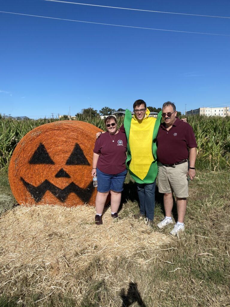 Student Chris Barron dressed in a corn suit standing with two other people in front of a hay bale painted to look like a pumpkin.
