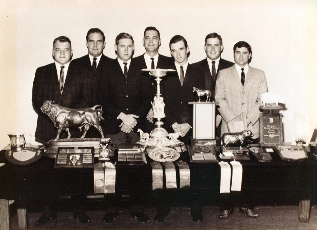 una foto más antigua en tonos sepia de hombres jóvenes con traje y corbata posando detrás de una serie de trofeos y cintas