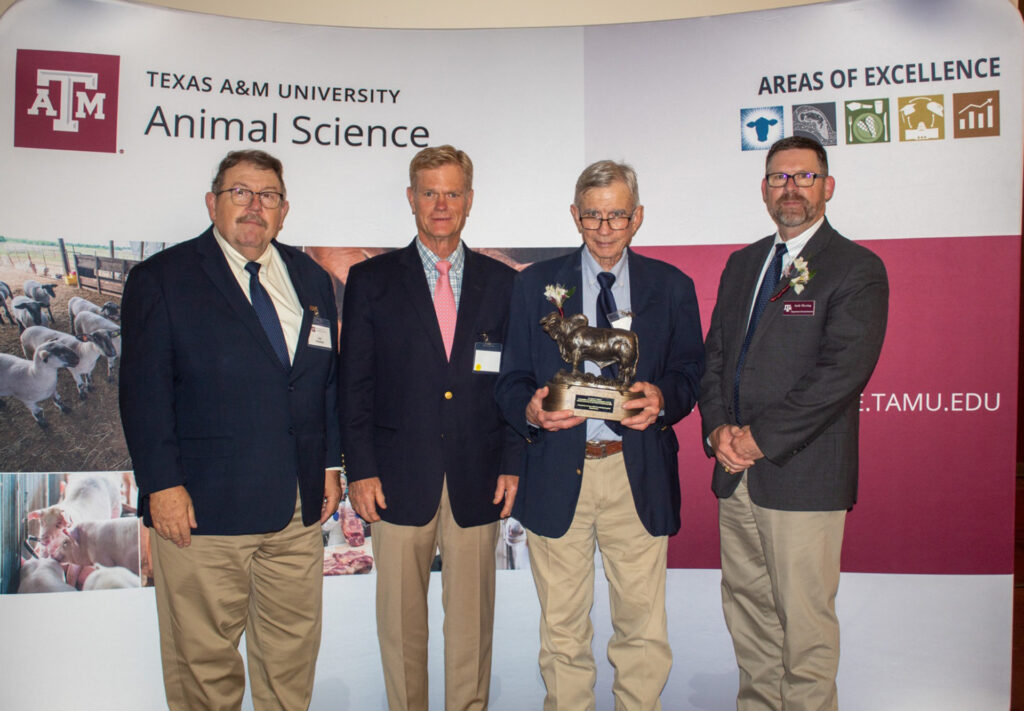 Cuatro hombres con traje y corbata posan frente a una pancarta de ciencia animal durante un programa de premios en el que Jim Sanders recibió una escultura de un toro.