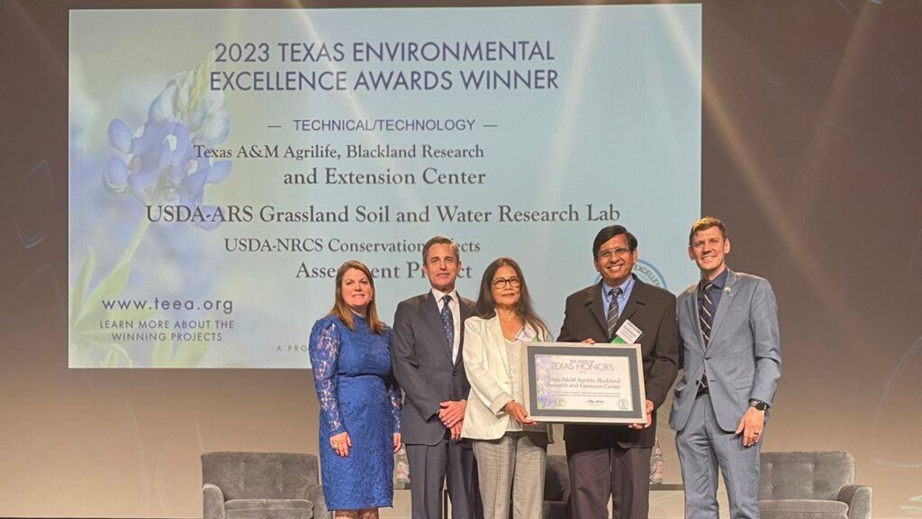 Five people in professional attire standing on stage holding a technology award certificate. Large screen in the background shows image of certificate.