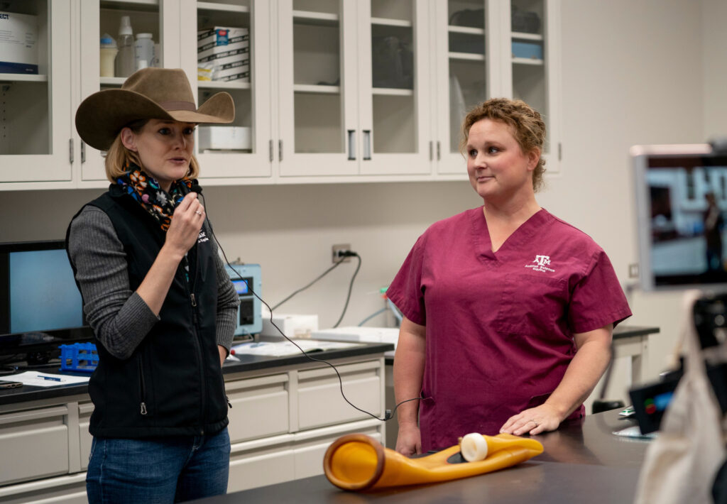 Una mujer con ropa vaquera y un sombrero de vaquero habla por un micrófono mientras otra mujer vestida con una bata médica observa.