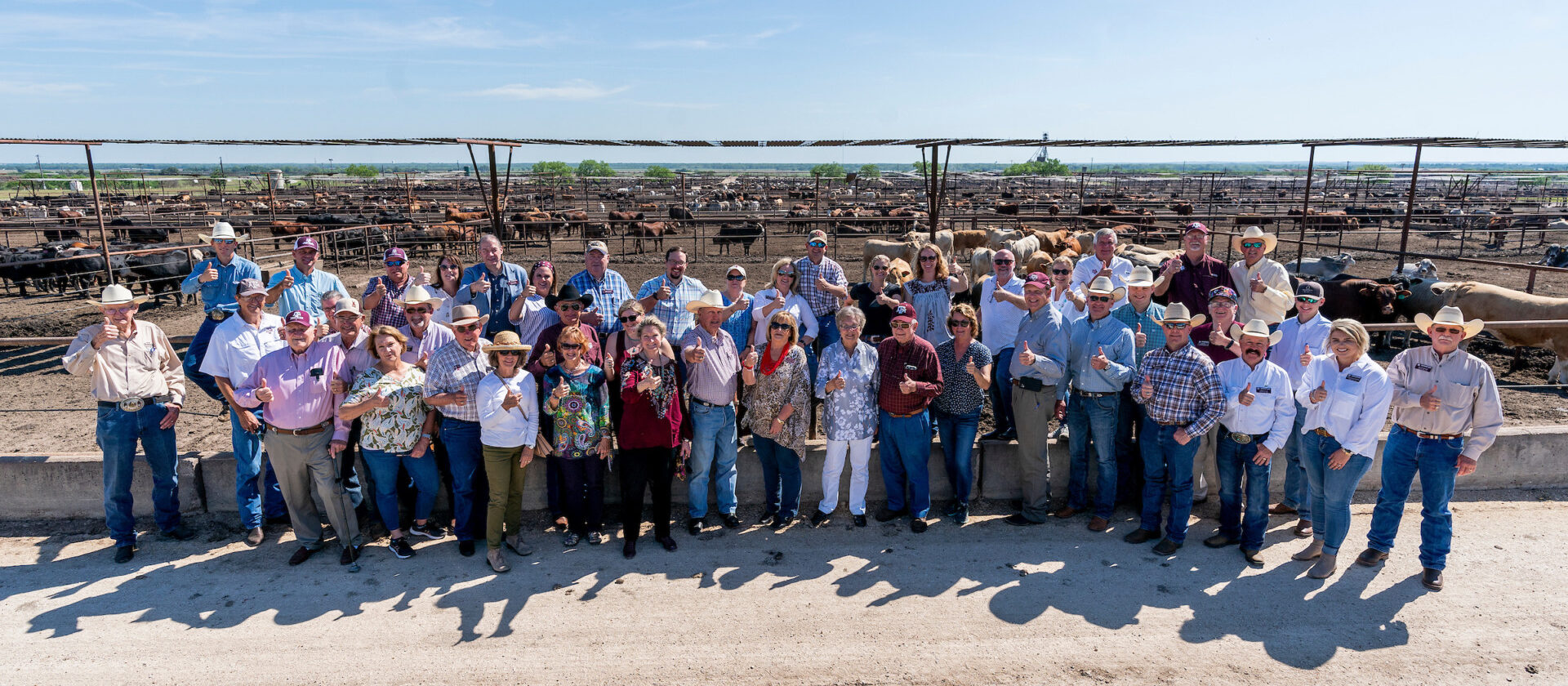 A group of men and women stand together in front of a feed yard with cattle in the background.