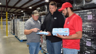 Three men stand in an egg warehouse looking at two cartons of eggs.